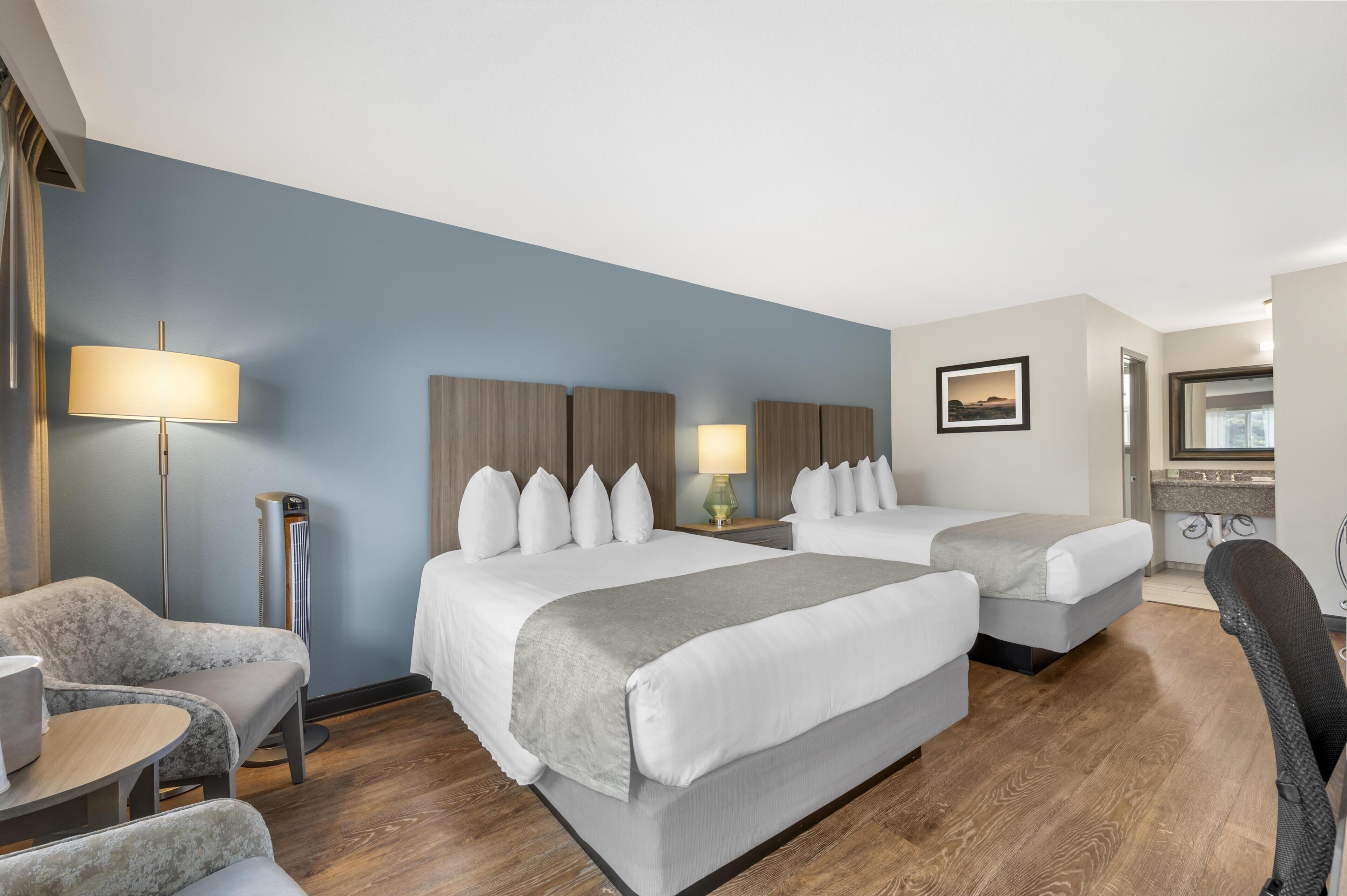 Ein Hotelzimmer mit zwei Betten, einem Schreibtisch, einem Stuhl, einer Stehlampe, einem Heizlüfter und einem Badezimmer mit Spiegel und Waschbecken.