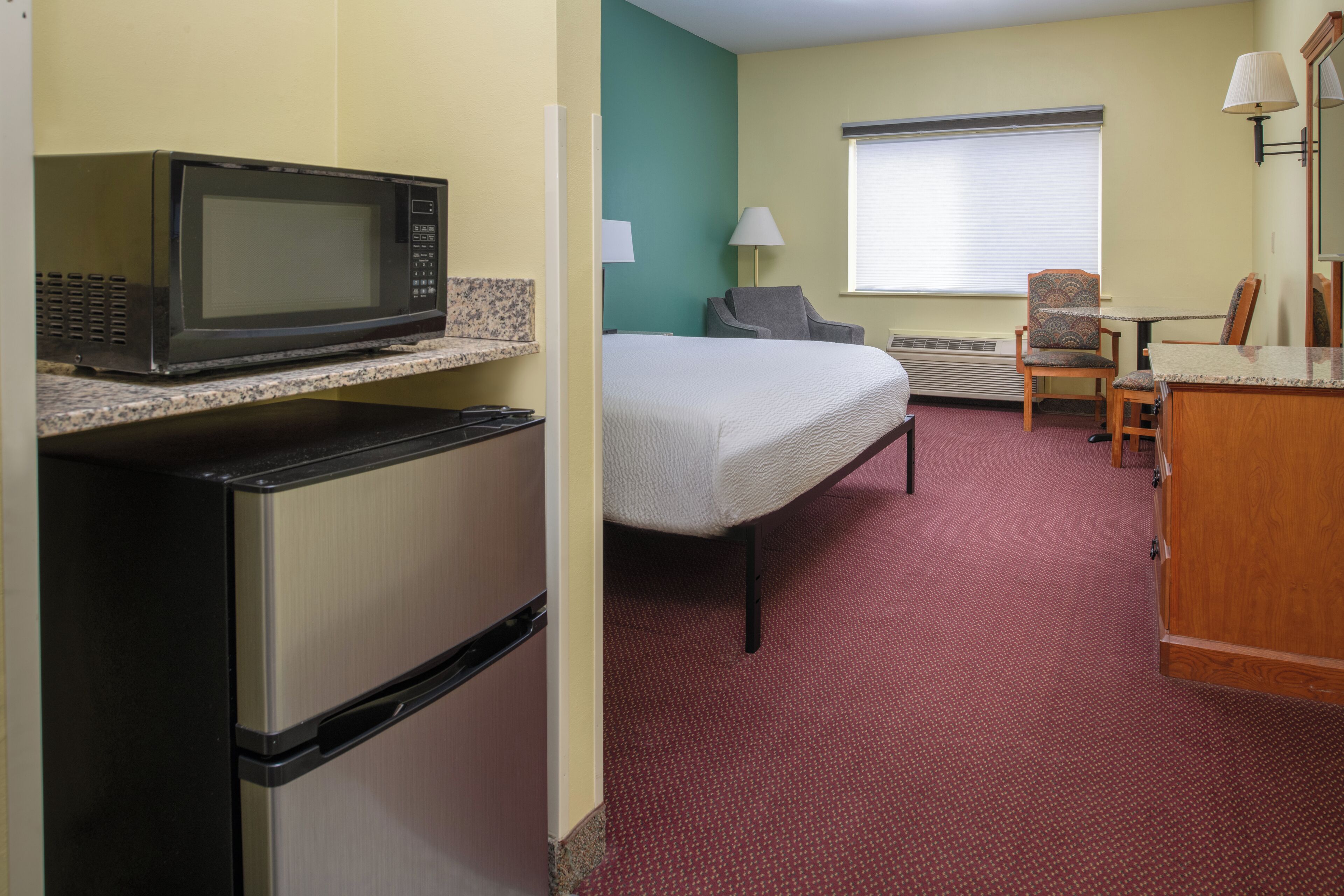 A hotel room with a bed, a microwave on a counter, a refrigerator, a chair, a table, and a window with blinds.