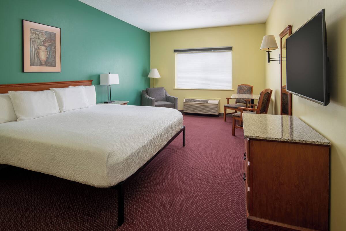 A hotel room with a large bed, a desk with a flat-screen TV, a chair, an air conditioning unit, and a window with blinds.