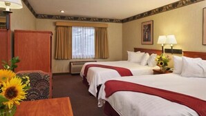 A hotel room with two beds, a wooden desk, a chair, a window with curtains, and a wall-mounted lamp. - Ashley Inn of Tillamook (Tillamook)