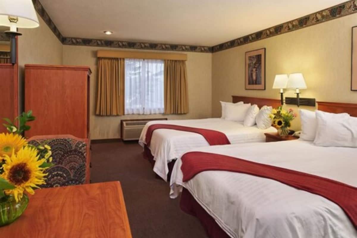 A hotel room with two beds, a wooden desk, a chair, a window with curtains, and a wall-mounted lamp.