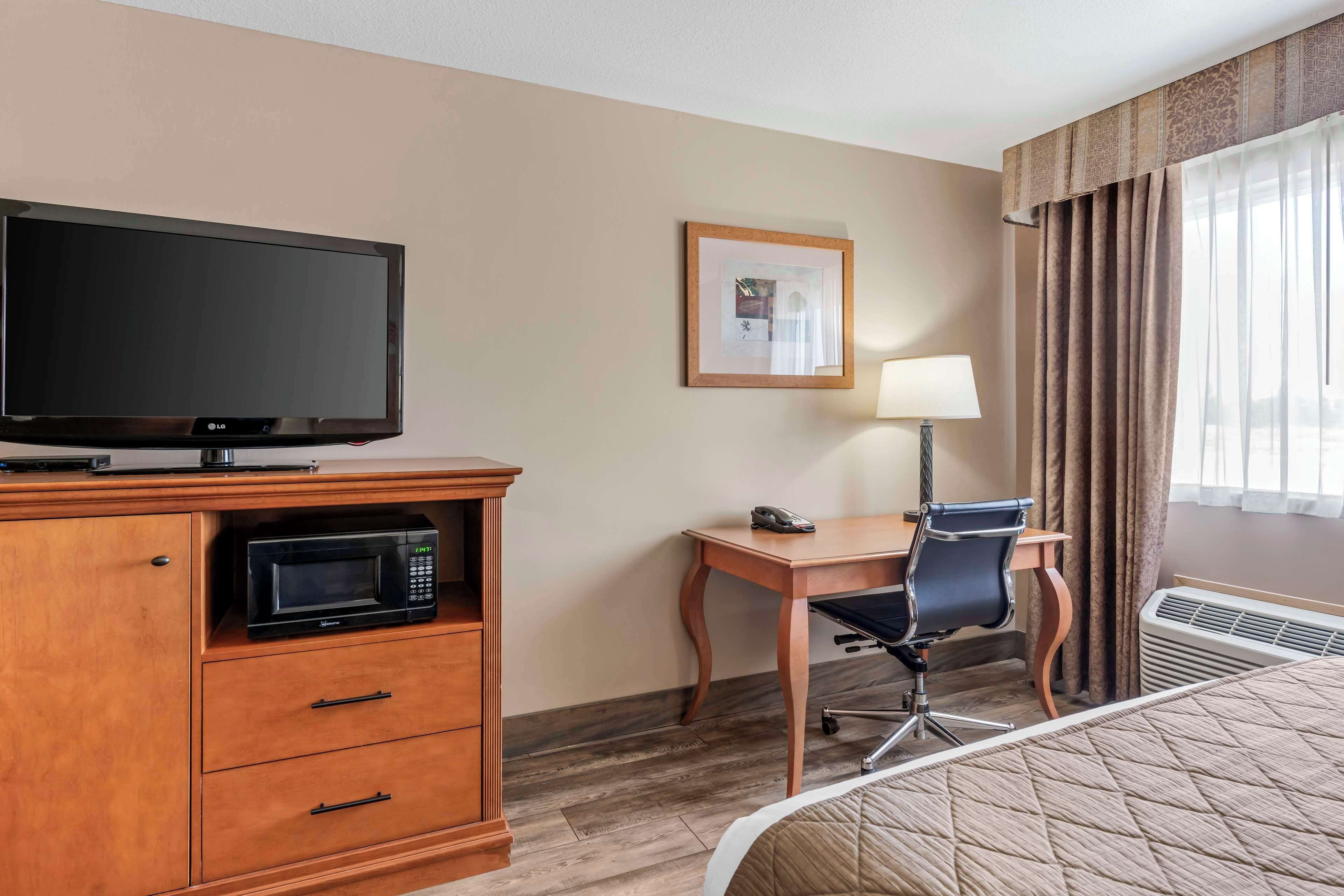 A hotel room with a flat-screen TV, a microwave, a desk with a chair, a framed picture on the wall, and a bed with a quilt.