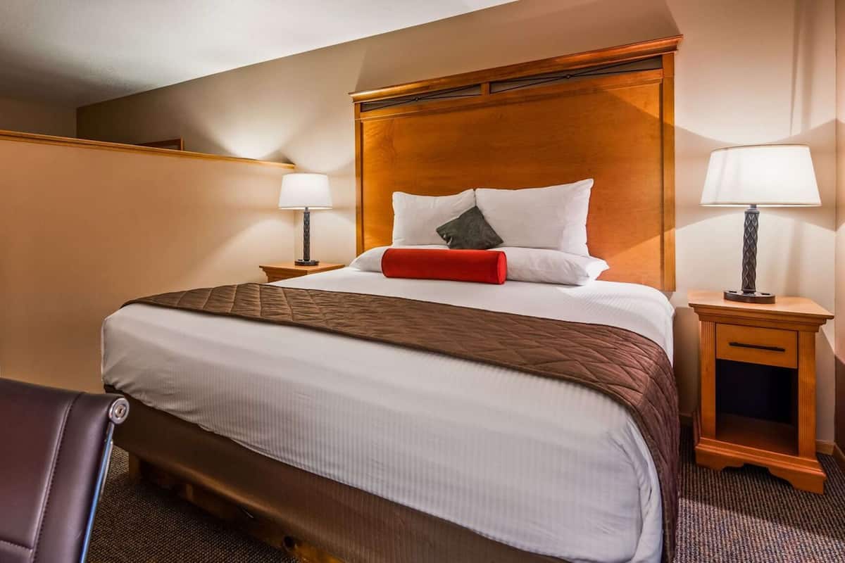 A hotel room with a large bed, two bedside tables with lamps, a wooden headboard, a red accent pillow, a brown quilted bedspread, a leather chair, and a carpeted floor.