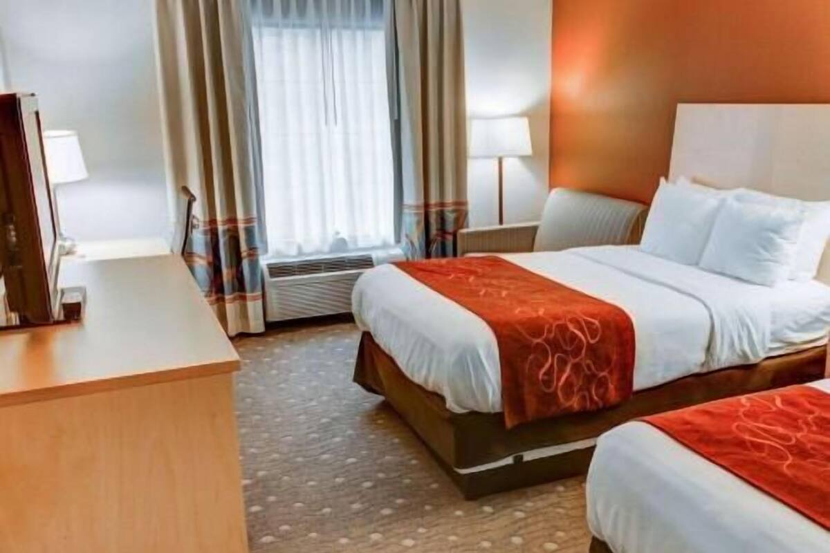 A hotel room with a bed, a desk, a chair, a window with curtains, and a wall-mounted lamp.