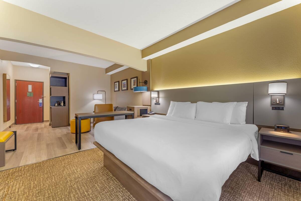 A hotel room with a large bed, a desk, a chair, a small kitchenette, and a bathroom door.