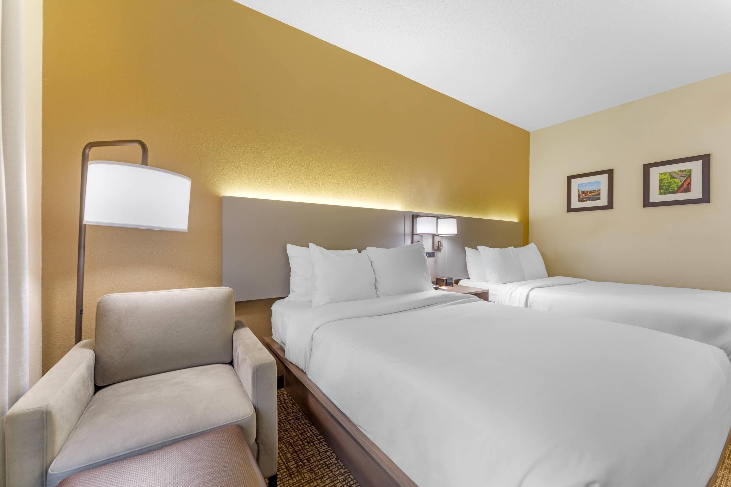 A hotel room with two beds, a chair, a lamp, and two framed pictures on the wall.