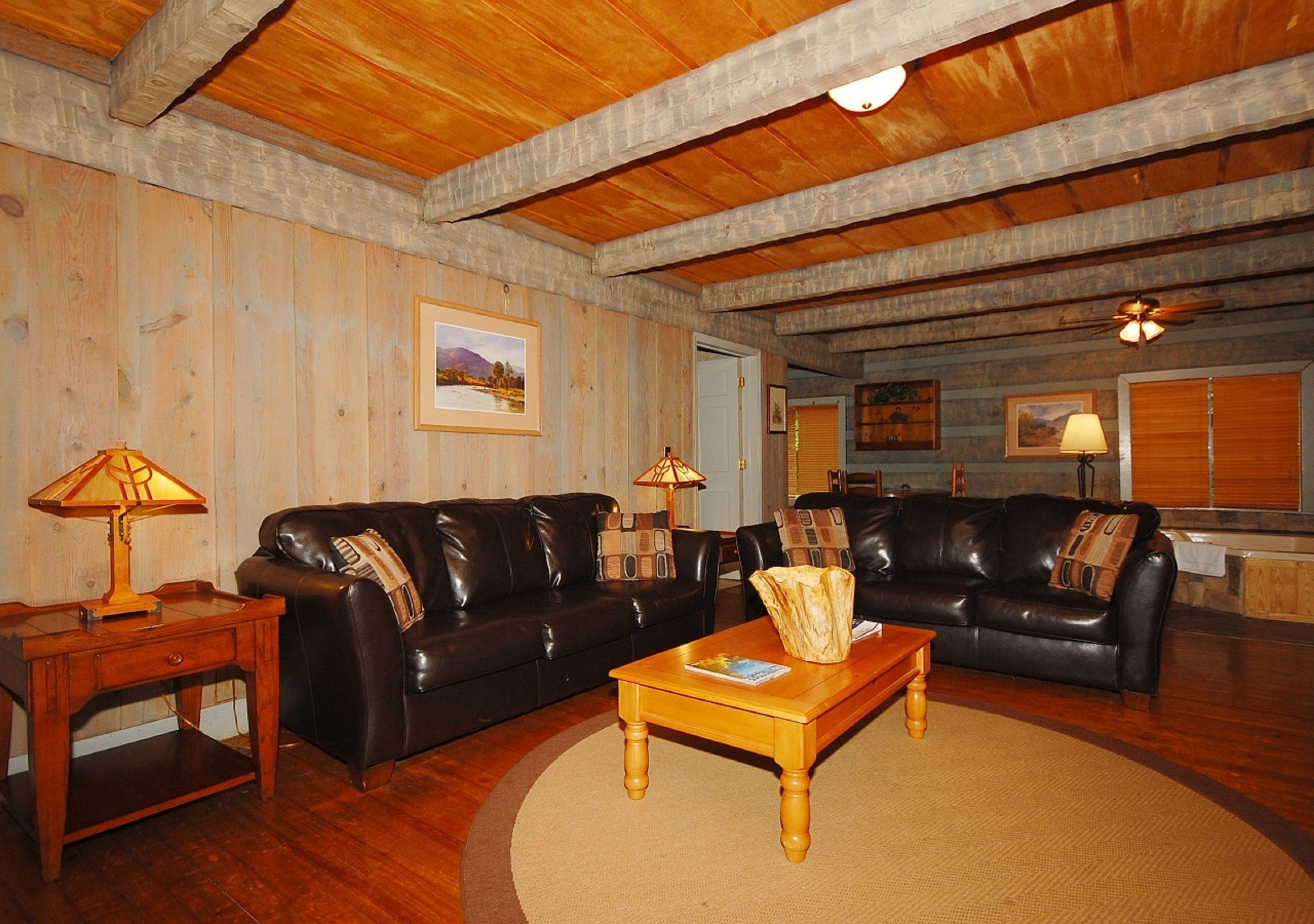 A cozy living room with wooden ceilings, two leather sofas, a wooden coffee table, and a small side table with a lamp.