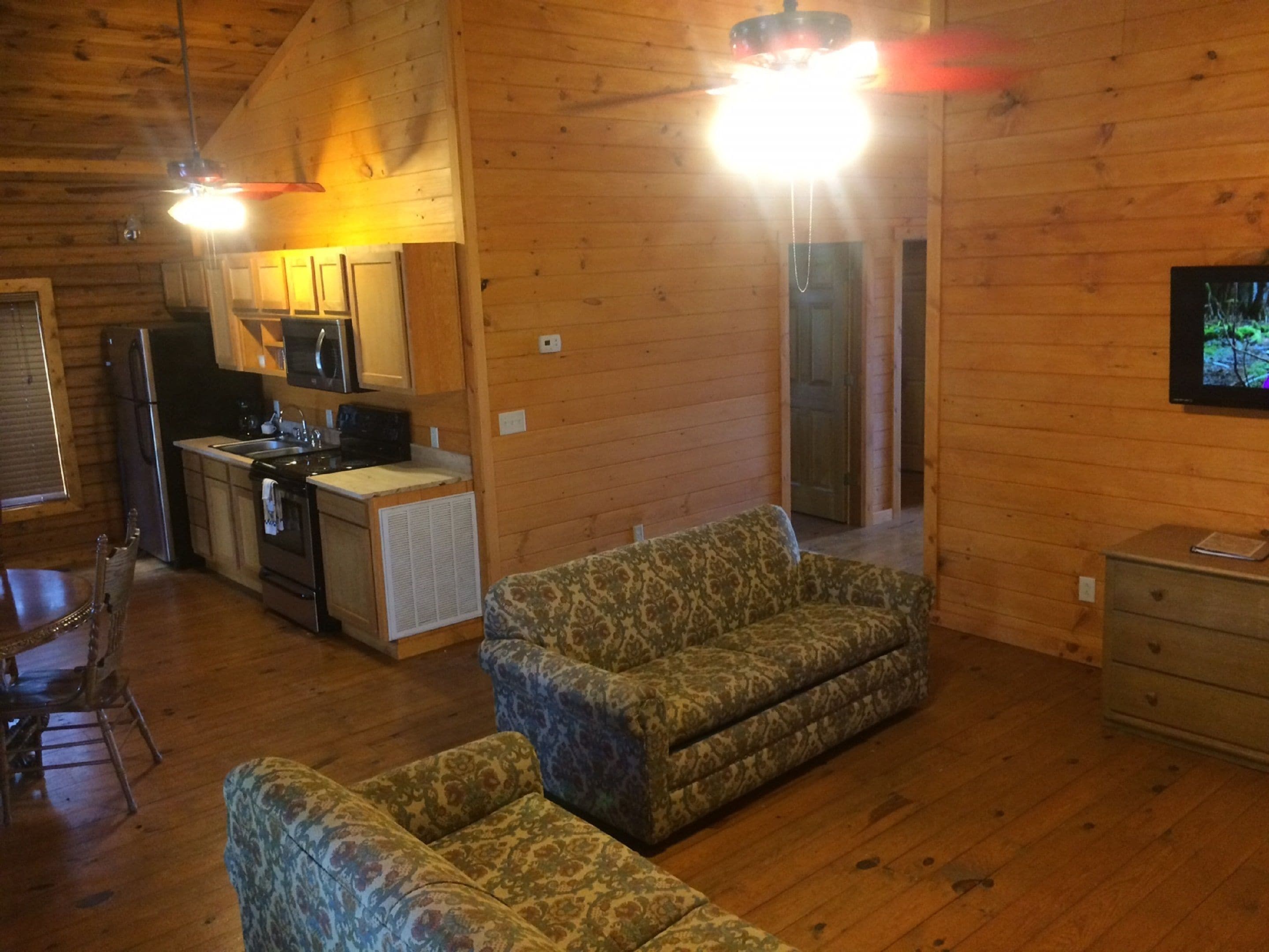 Un interior de cabaña acogedor con piso de madera, un sofá, una mesa de comedor con sillas, una zona de cocina con refrigerador, estufa y microondas, y un televisor montado en la pared.