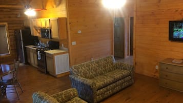 Un interior de cabaña acogedor con piso de madera, un sofá, una mesa de comedor con sillas, una zona de cocina con refrigerador, estufa y microondas, y un televisor montado en la pared.