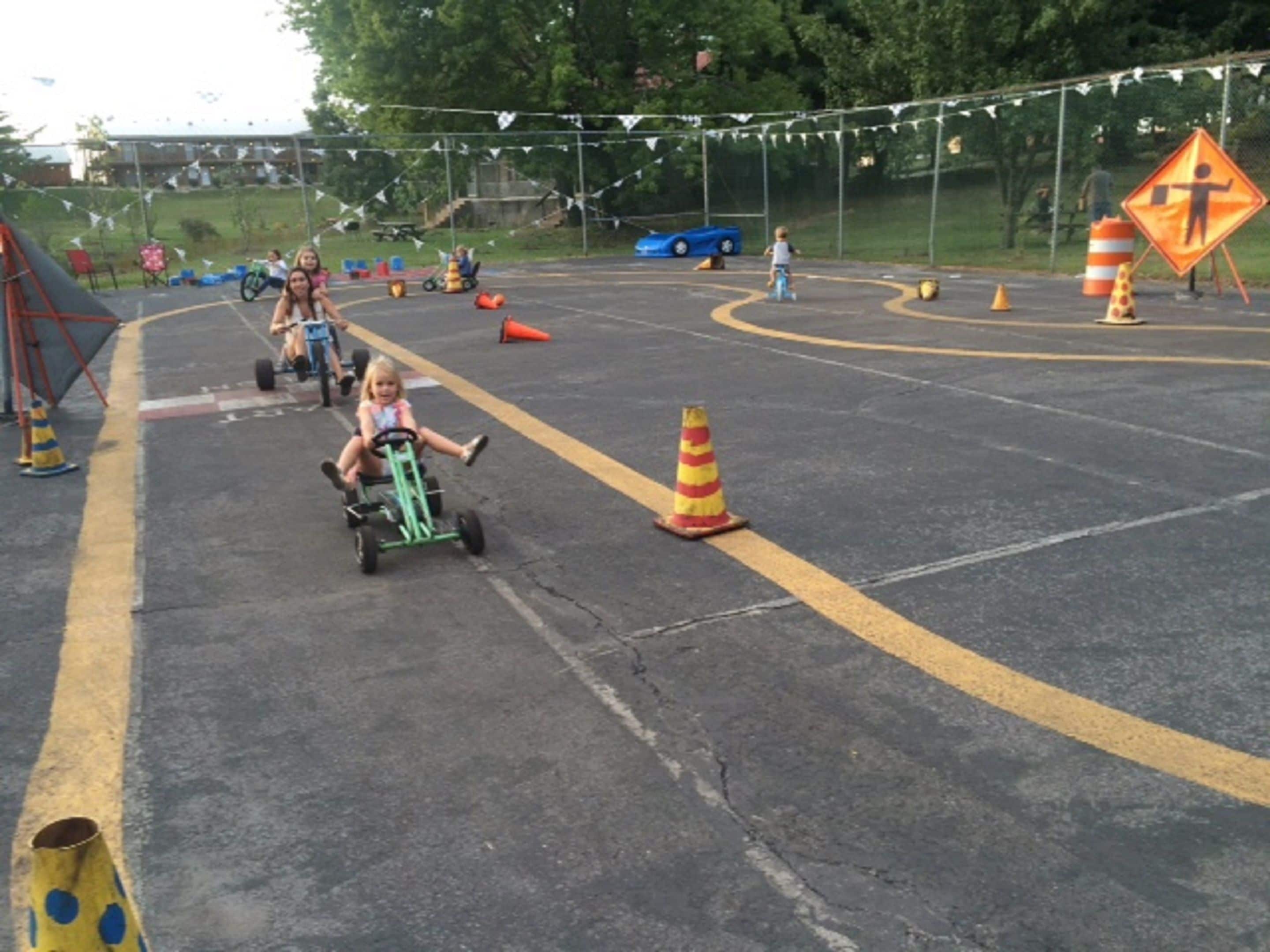 A parking lot with a child riding a tricycle, surrounded by traffic cones and a blue car in the background.