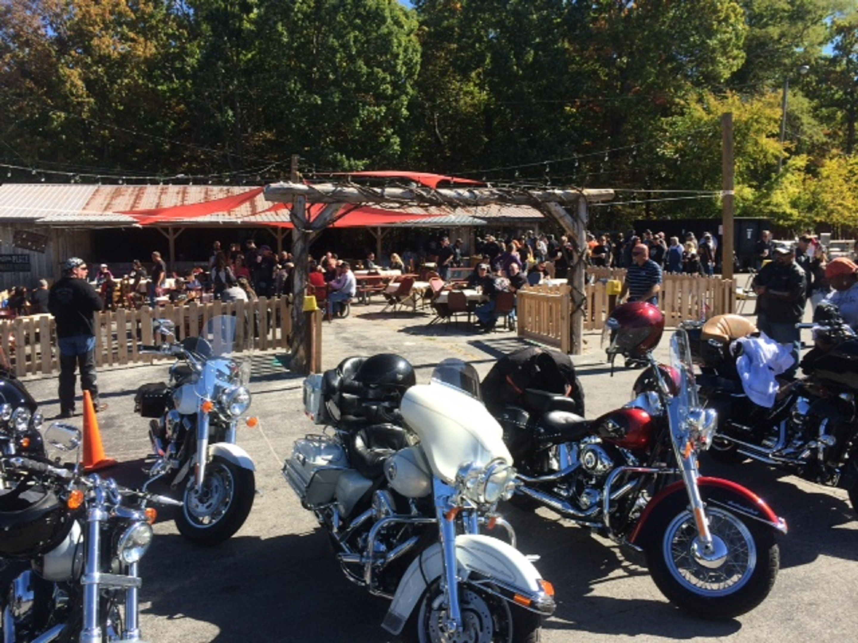 A parking area with several motorcycles lined up, a covered seating area with tables and chairs, and a crowd of people in the background.