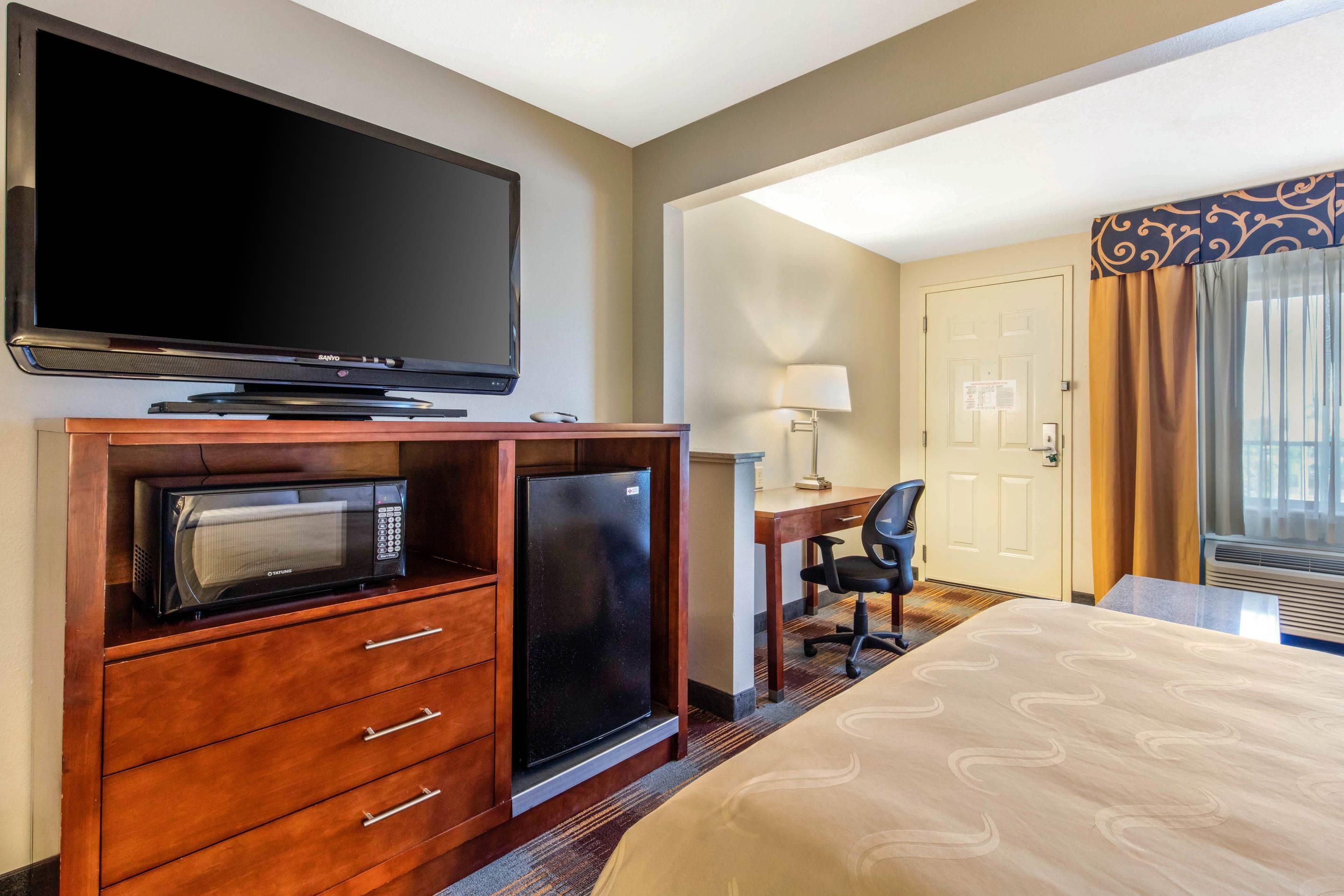 A hotel room with a flat-screen TV, a microwave, a refrigerator, a desk with a chair, and a bed with a patterned bedspread.