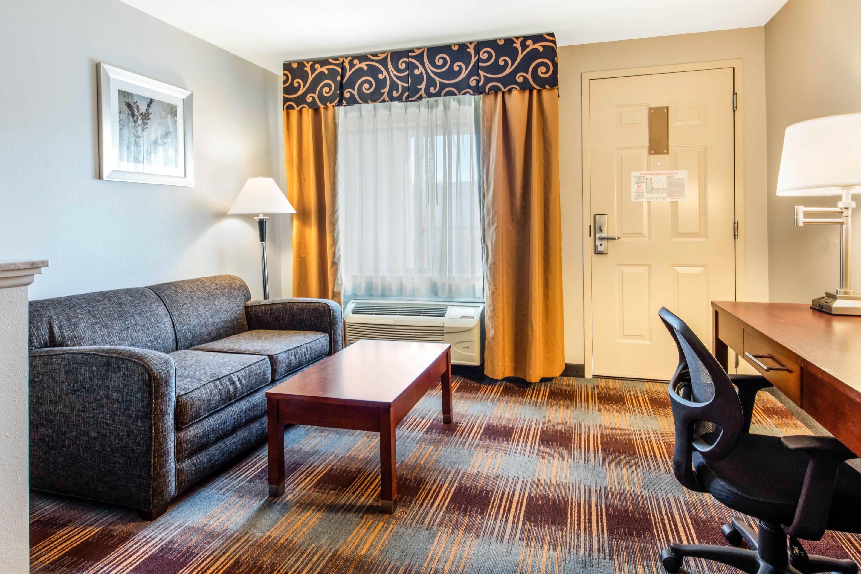 Habitación de hotel con sofá, una mesita, escritorio con silla, ventana con cortinas, equipo de aire acondicionado y puerta con plano de evacuación.