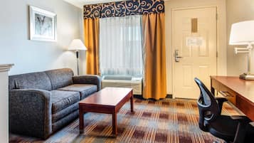 Habitación de hotel con sofá, una mesita, escritorio con silla, ventana con cortinas, equipo de aire acondicionado y puerta con plano de evacuación.