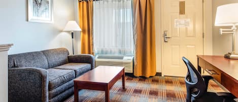 Habitación de hotel con sofá, una mesita, escritorio con silla, ventana con cortinas, equipo de aire acondicionado y puerta con plano de evacuación.