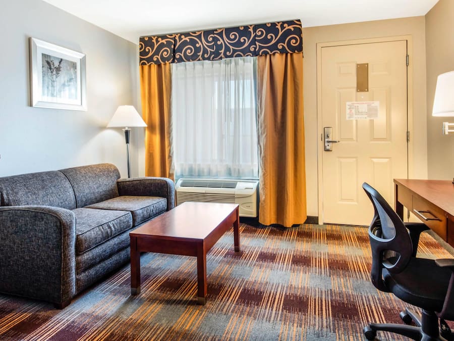 Habitación de hotel con sofá, una mesita, escritorio con silla, ventana con cortinas, equipo de aire acondicionado y puerta con plano de evacuación.