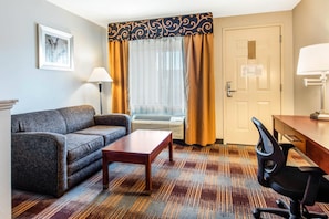 A hotel room with a sofa, a small table, a desk with a chair, a window with curtains, an air conditioning unit, and a door with an emergency exit plan. - Quality Inn Memphis Airport (Memphis)