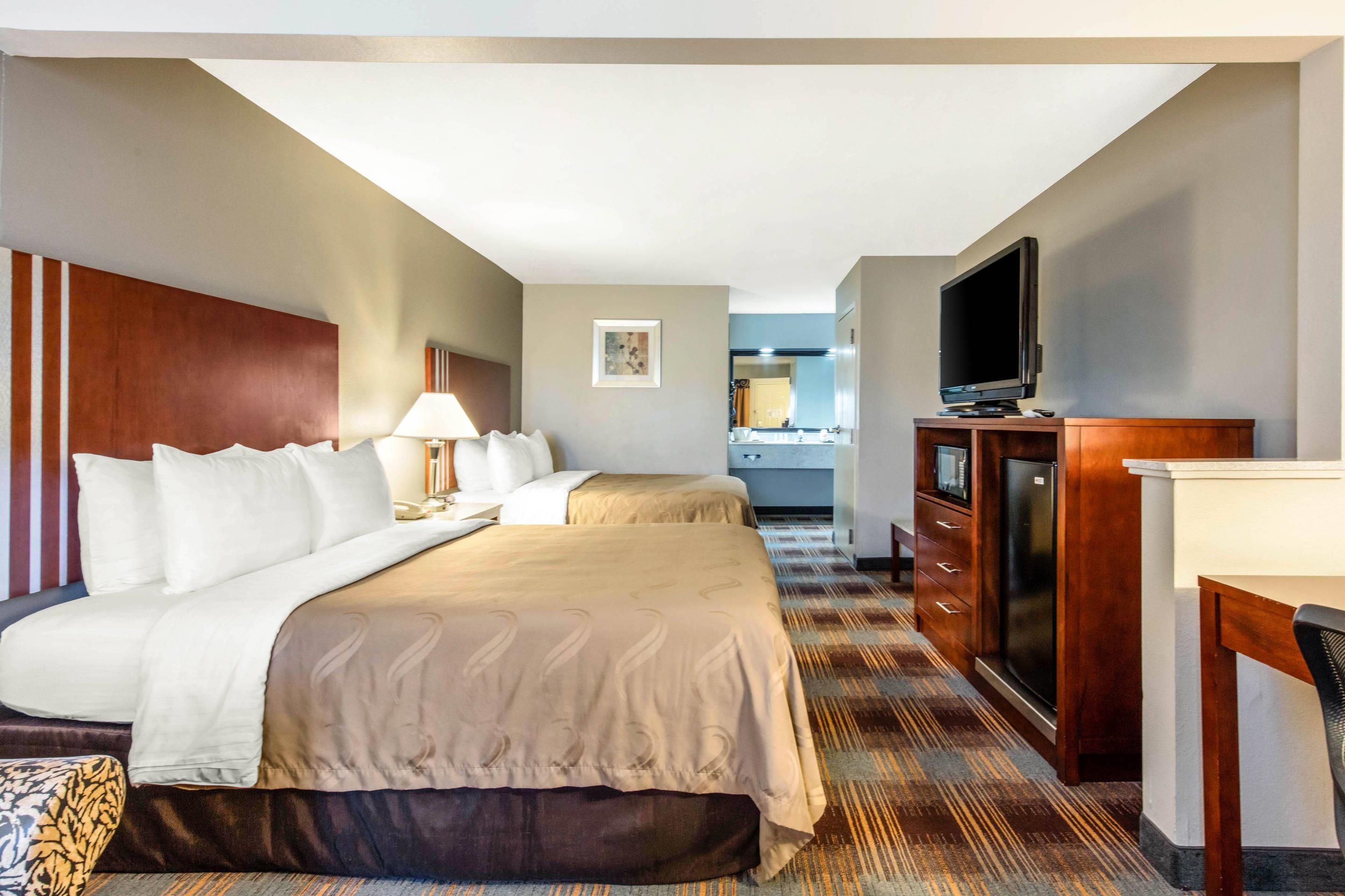 A hotel room with two beds, a desk, a television, and a bathroom visible in the background.