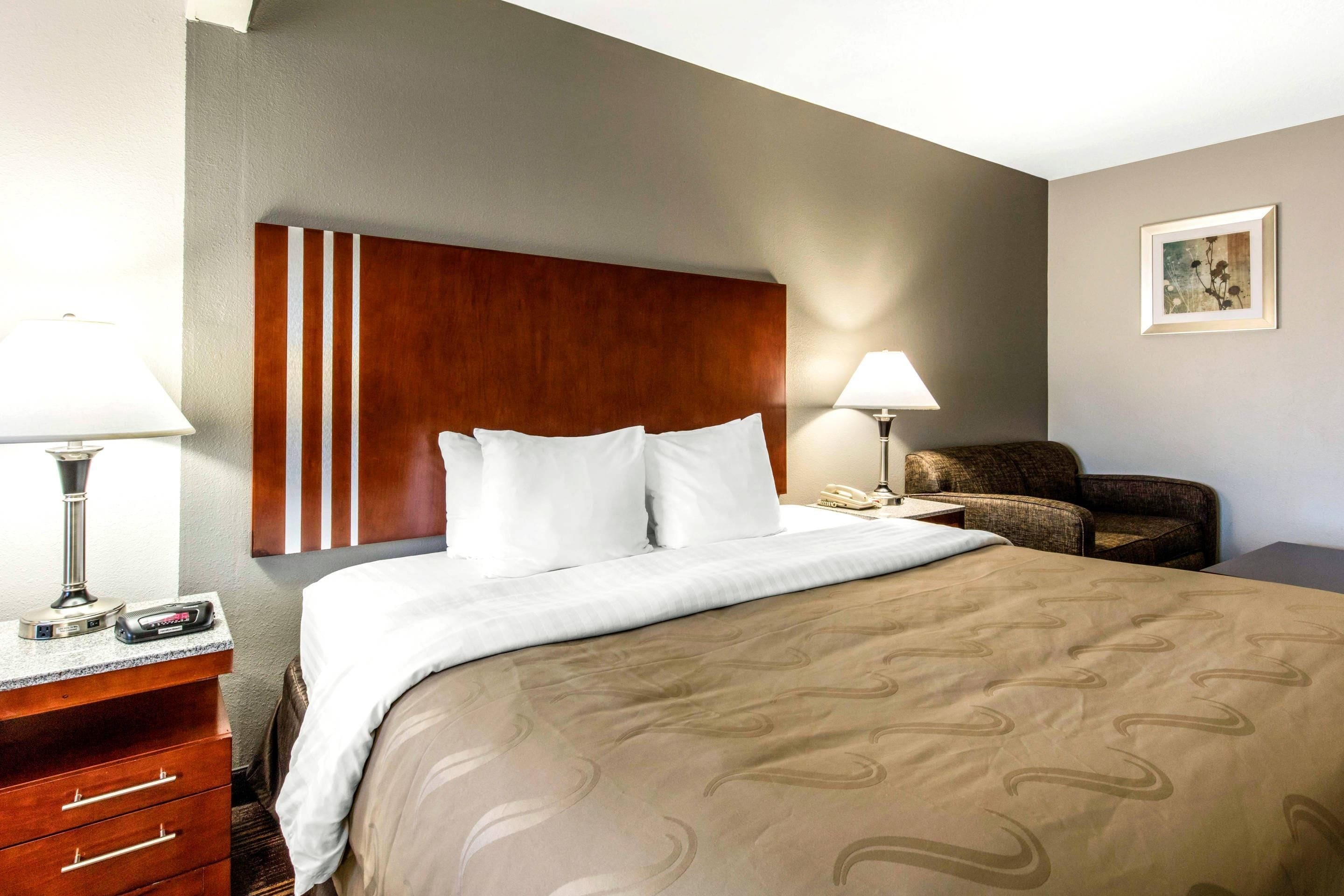 Habitación de hotel con cama, escritorio con lámpara, silla y mesita de noche con teléfono.
