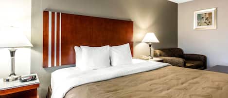 A hotel room with a bed, a desk with a lamp, a chair, and a bedside table with a phone.
