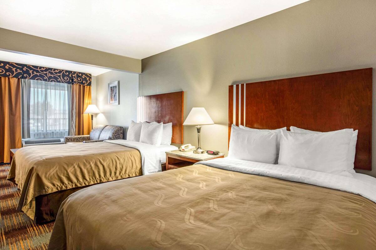 Habitación de hotel con dos camas, una mesita de noche con lámpara, teléfono y ventana con cortinas.