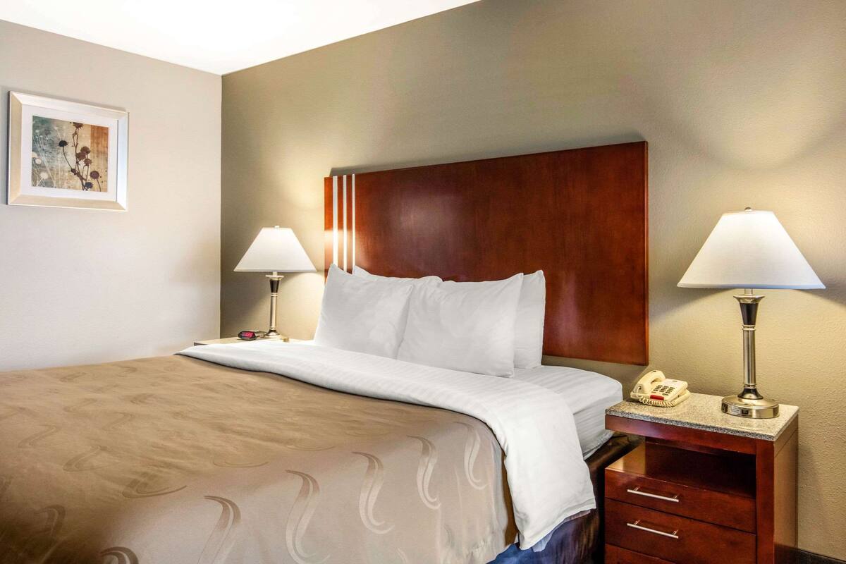 Habitación de hotel con una cama grande, dos lámparas de noche, un cabecero de madera, un cuadro en la pared y un teléfono en la mesita de noche.