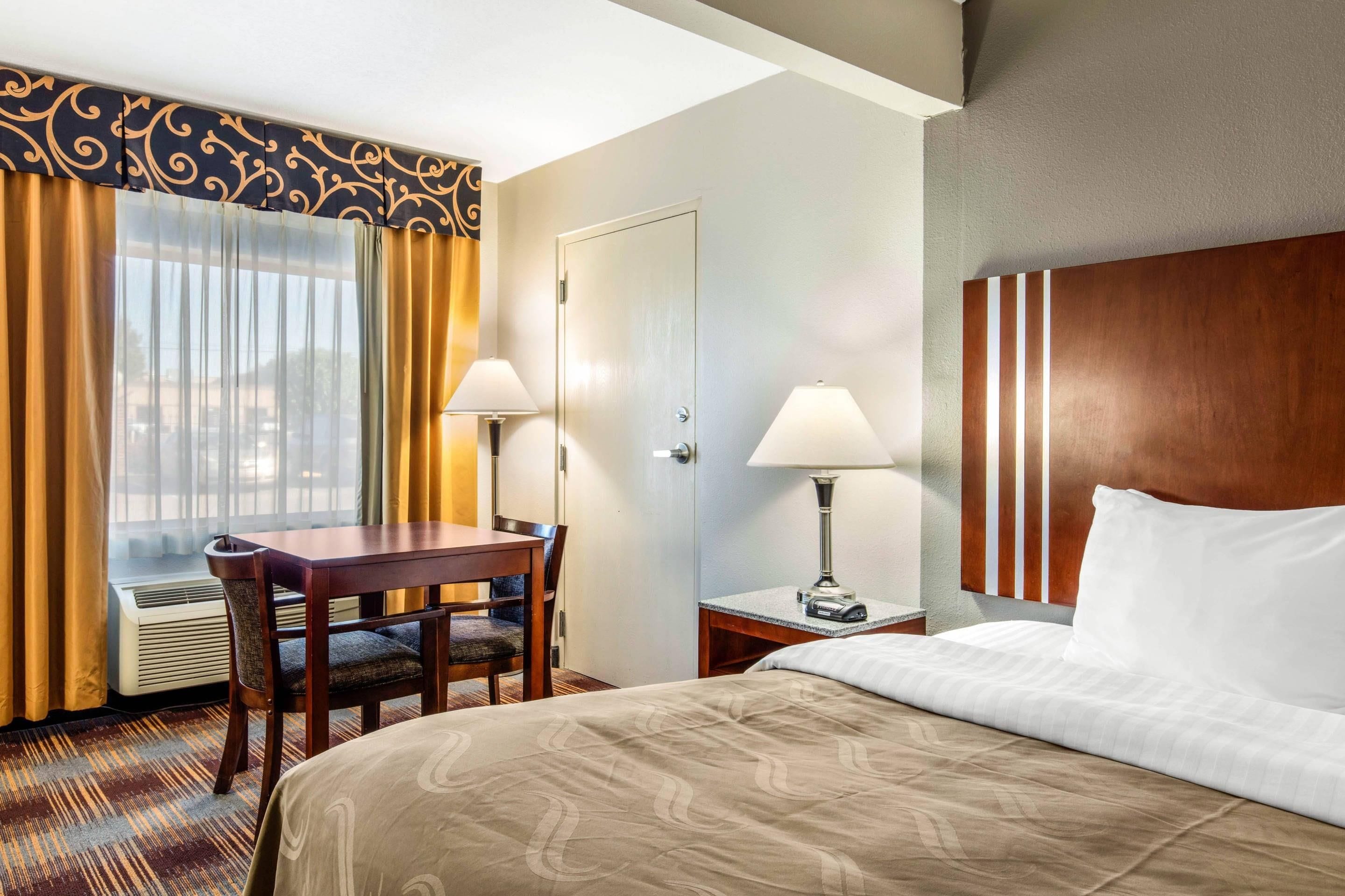 A hotel room with a bed, a desk, two chairs, a lamp, a mirror, and a window with curtains.