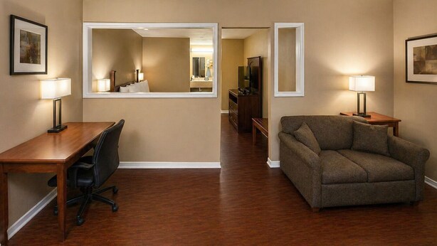 A hotel room with a wooden desk, a black office chair, a brown sofa, two bedside lamps, a large mirror, and a television.
