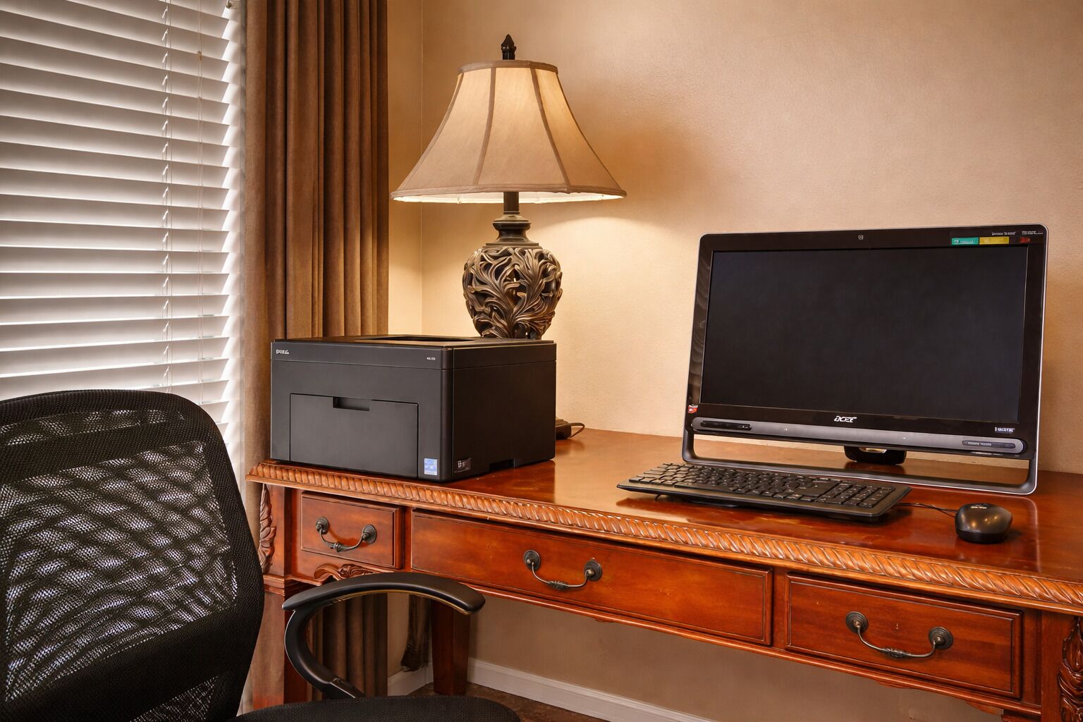 A hotel room with a wooden desk, a desktop computer, a lamp, and a chair.