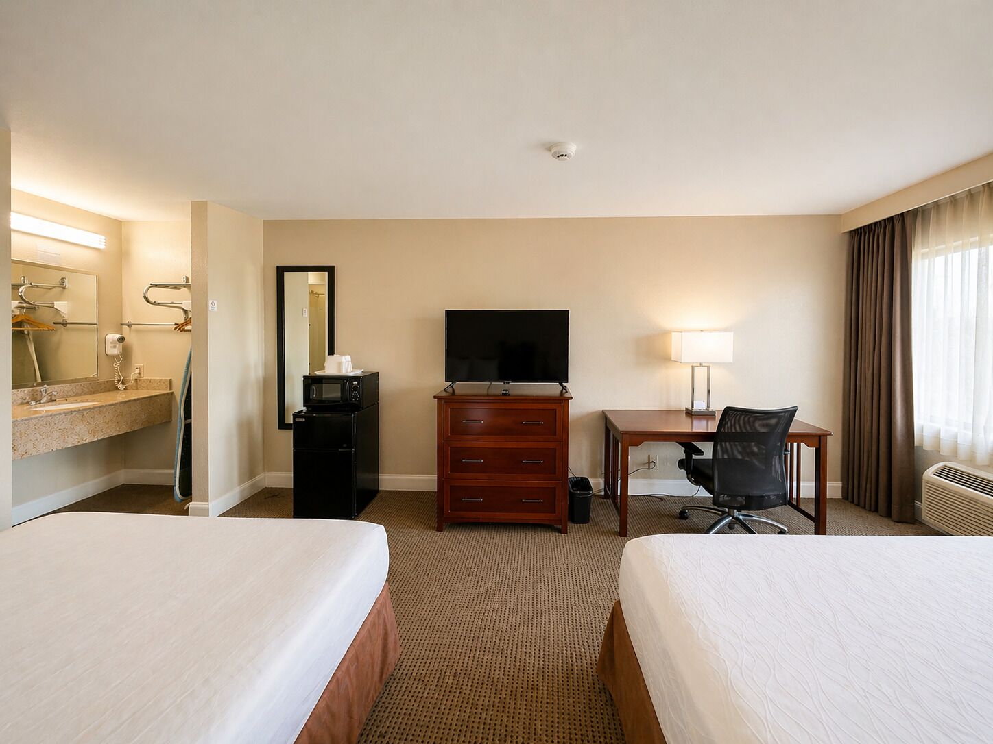 Habitación de hotel con dos camas, un escritorio con silla, televisor, espejo, cómoda y baño con lavamanos y espejo.