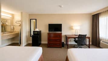 Habitación de hotel con dos camas, un escritorio con silla, televisor, espejo, cómoda y baño con lavamanos y espejo.