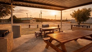 An outdoor patio with picnic tables and a barbecue grill, surrounded by a concrete floor and a metal roof structure.