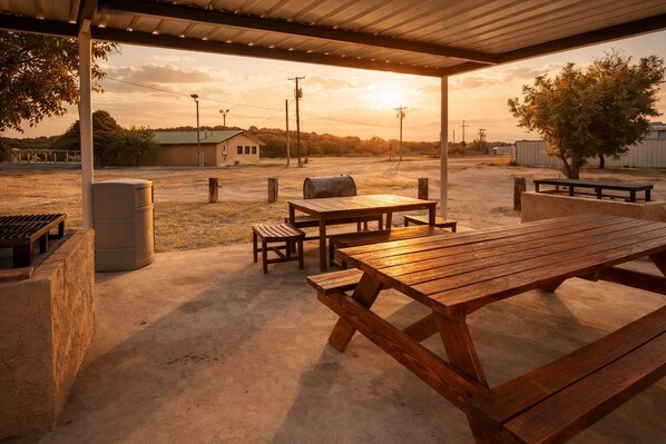 An outdoor patio with picnic tables and a barbecue grill, surrounded by a concrete floor and a metal roof structure.