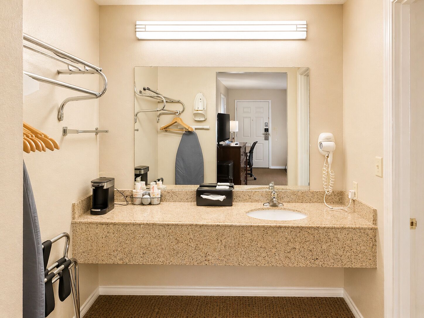 A hotel bathroom with a granite countertop, a sink, a mirror, a hairdryer, and a towel rack.