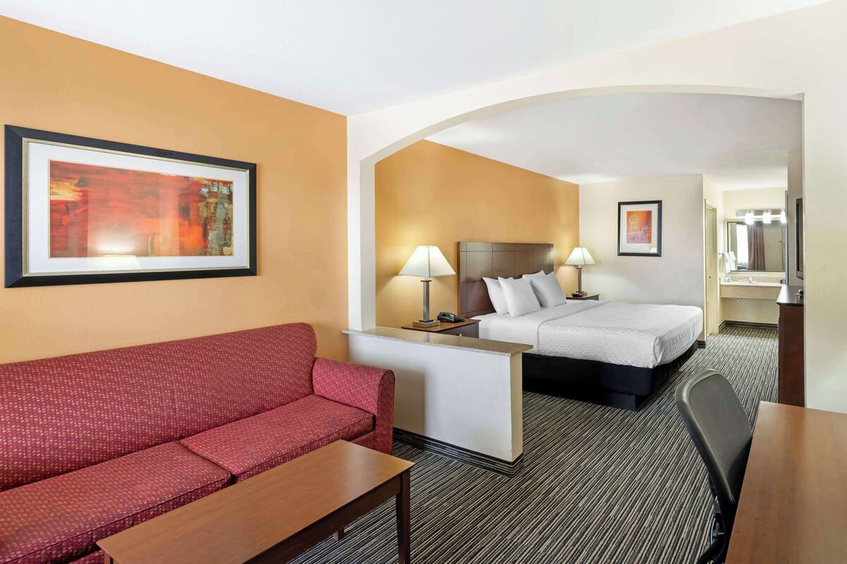 A hotel room with a red sofa, a bed with white bedding, a desk with a lamp, and a framed picture on the wall.
