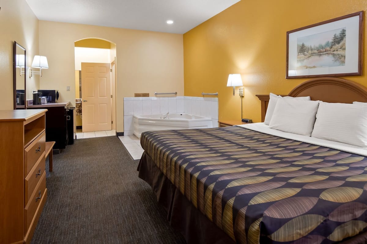 A hotel room with a bed, a desk, a chair, a TV, a bathroom with a bathtub, and a painting on the wall.