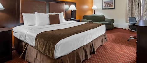 A hotel room with a large bed, two bedside tables with lamps, a green armchair, a desk with a chair, and a framed picture on the wall.
