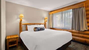 Cama bem arrumada com lençóis brancos, cabeceira de madeira, duas luminárias de cabeceira e janela com cortinas.