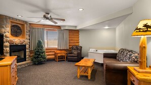 A cozy log cabin interior with a fireplace, a ceiling fan, a wooden coffee table, a brown leather sofa, and a patterned throw pillow.