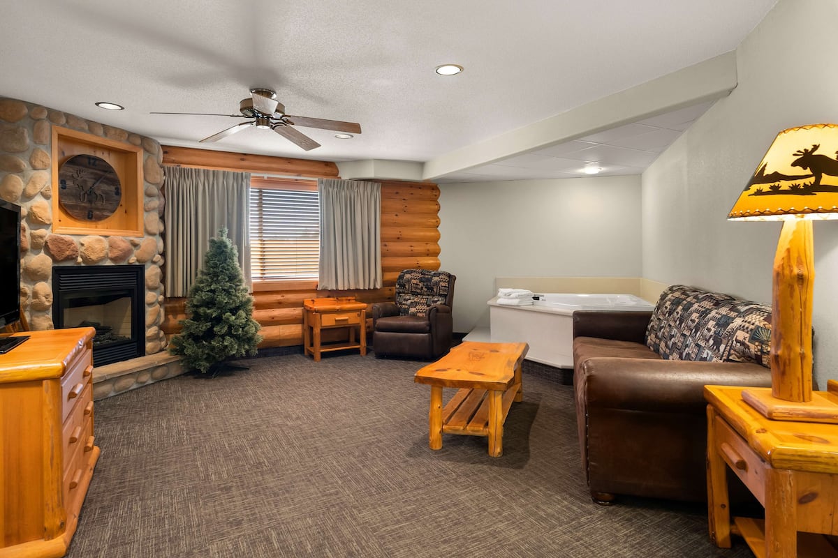 A cozy log cabin interior with a fireplace, a ceiling fan, a wooden coffee table, a brown leather sofa, and a patterned throw pillow.