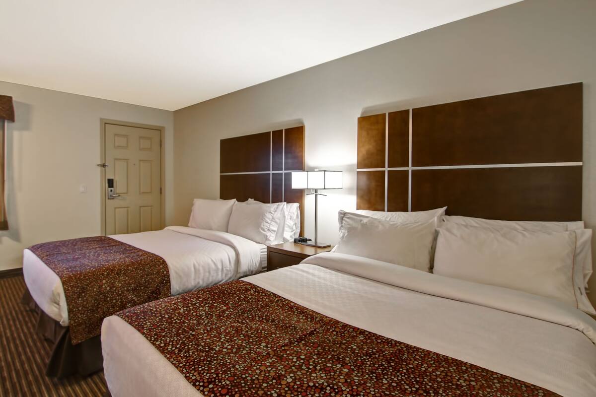 A hotel room with two beds, a door, a nightstand with a lamp, and a large window.