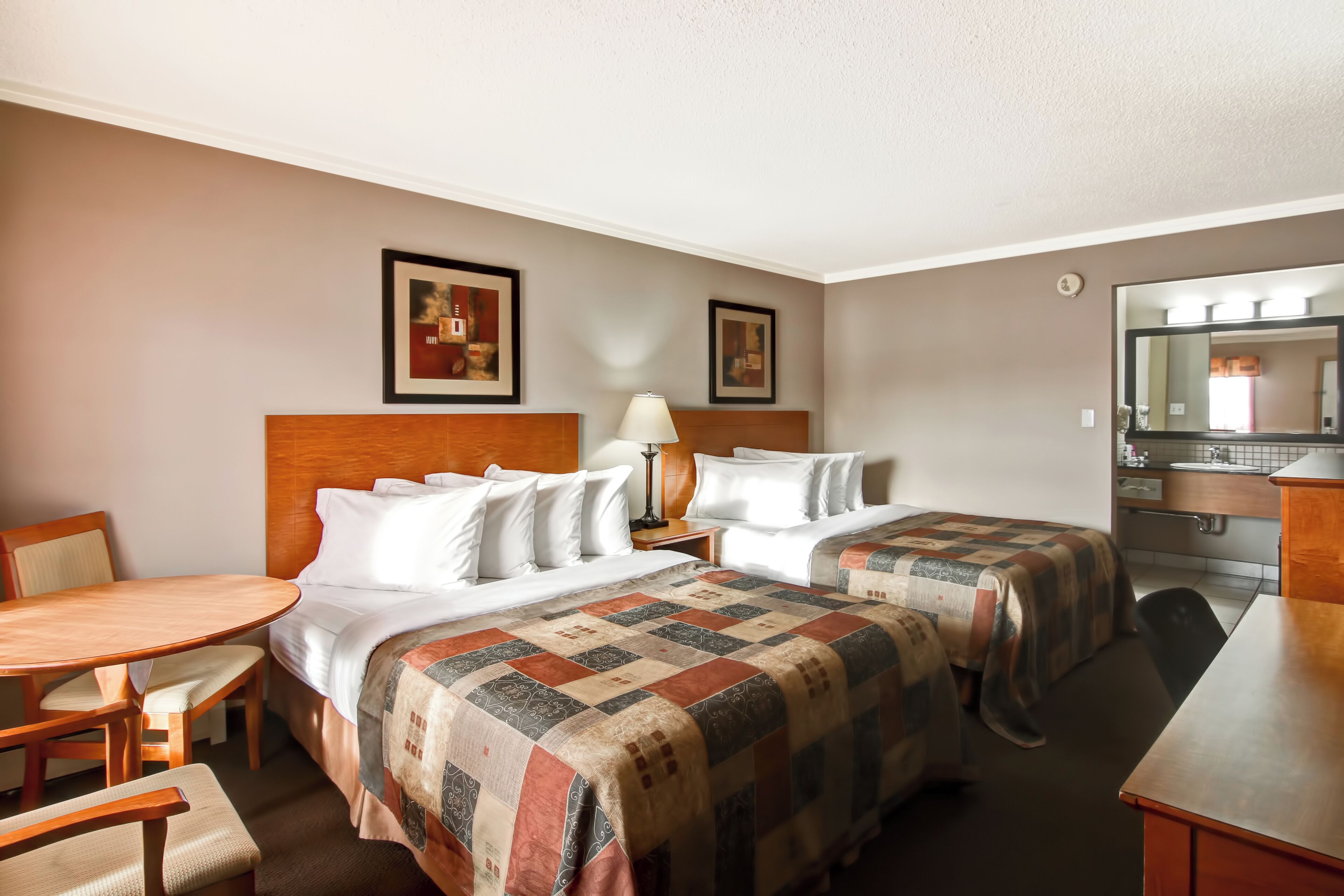 A hotel room with two beds, a wooden headboard, a round dining table, a small desk, and a mirror.
