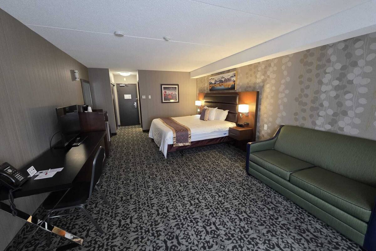 A hotel room with a bed, a desk, a green sofa, a painting on the wall, and a patterned carpet.