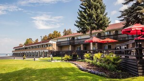 A hotel with a waterfront view, outdoor seating with red umbrellas, and a well-maintained lawn. - Ramada by Wyndham Jacksons Point (Georgina)