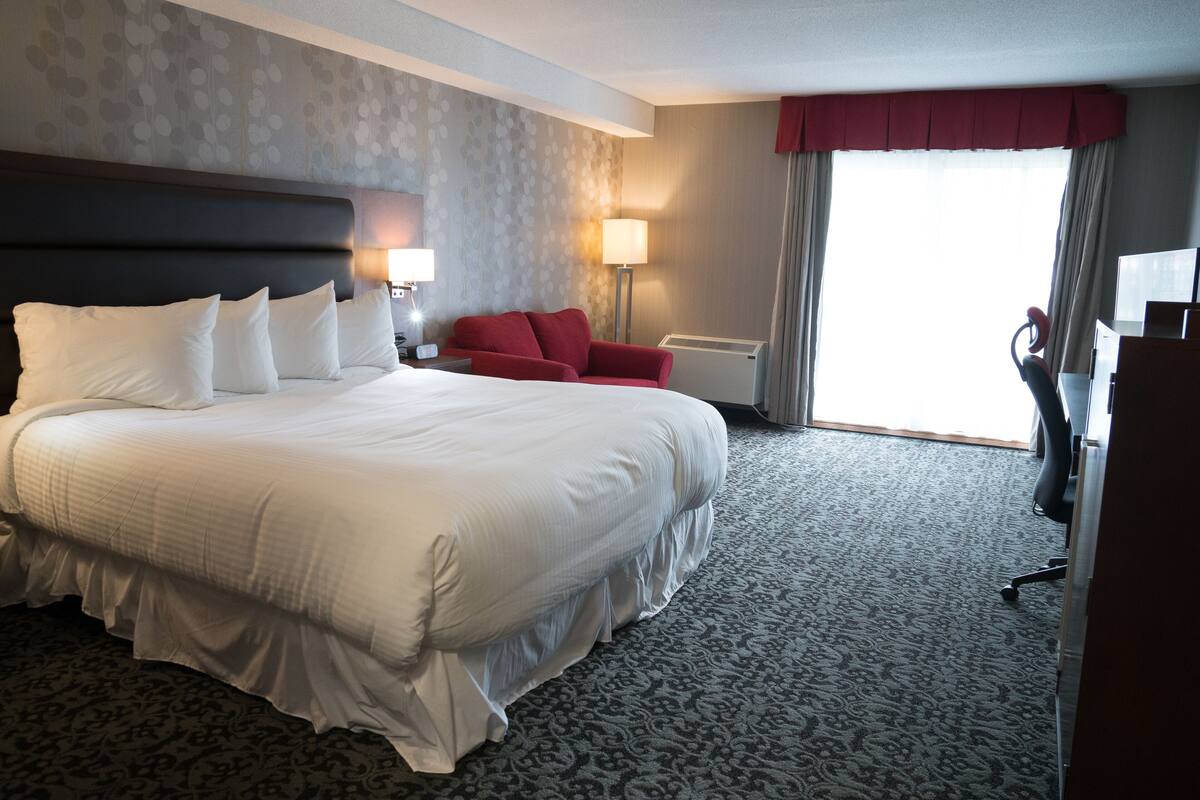 A hotel room with a large bed, a red armchair, a desk, and a view of the outdoors.