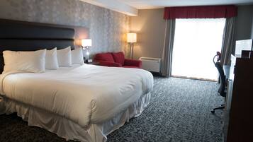 Quarto de hotel com uma cama grande, uma poltrona vermelha, uma mesa de trabalho e vista para o exterior.