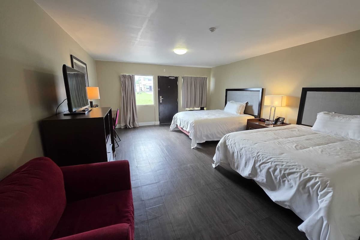 A hotel room with two beds, a desk with a TV, a red chair, and a door leading outside.