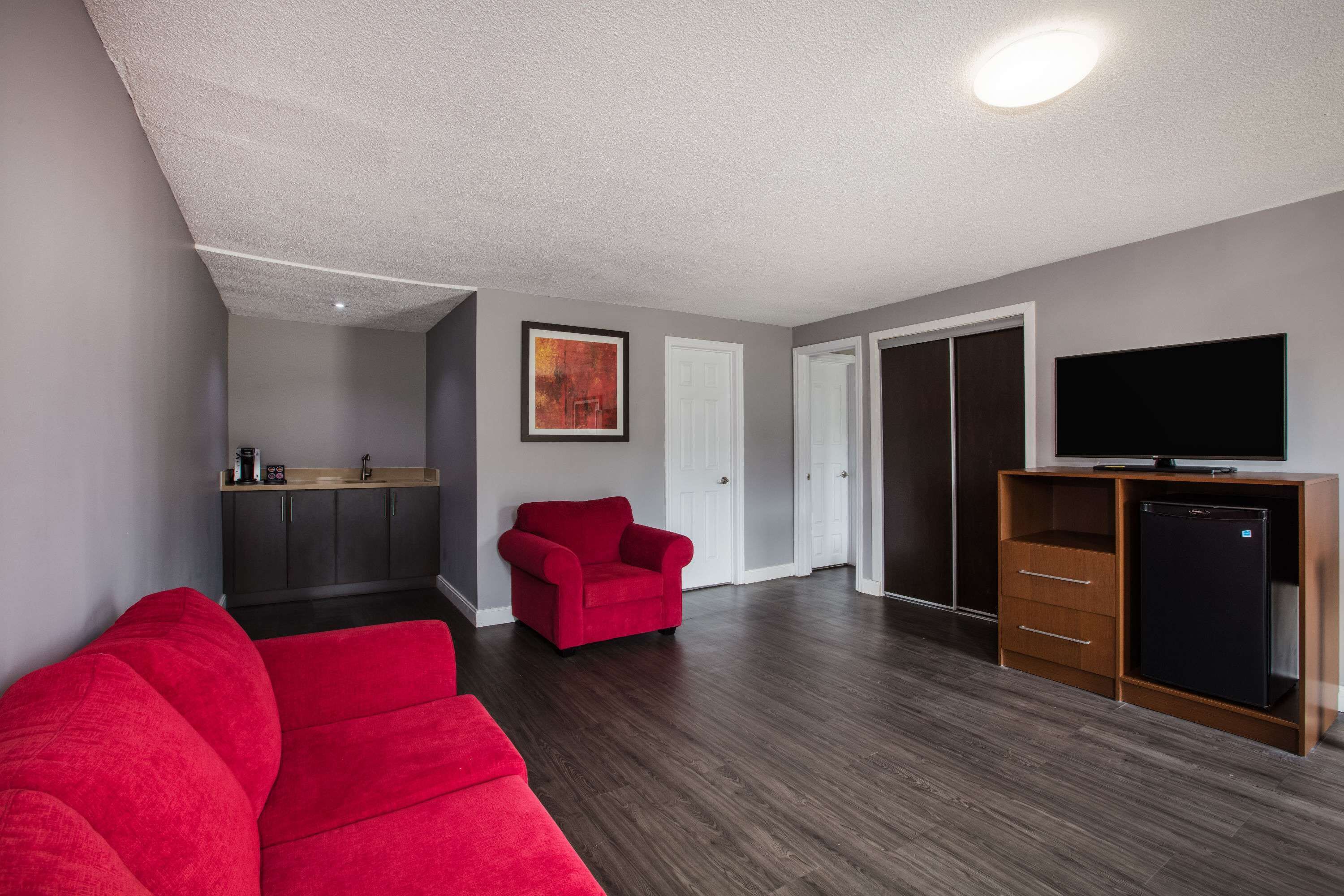 Una sala de estar con un sofá rojo, un sillón rojo, un televisor sobre un mueble de madera, y una zona de cocina con armarios y una encimera.