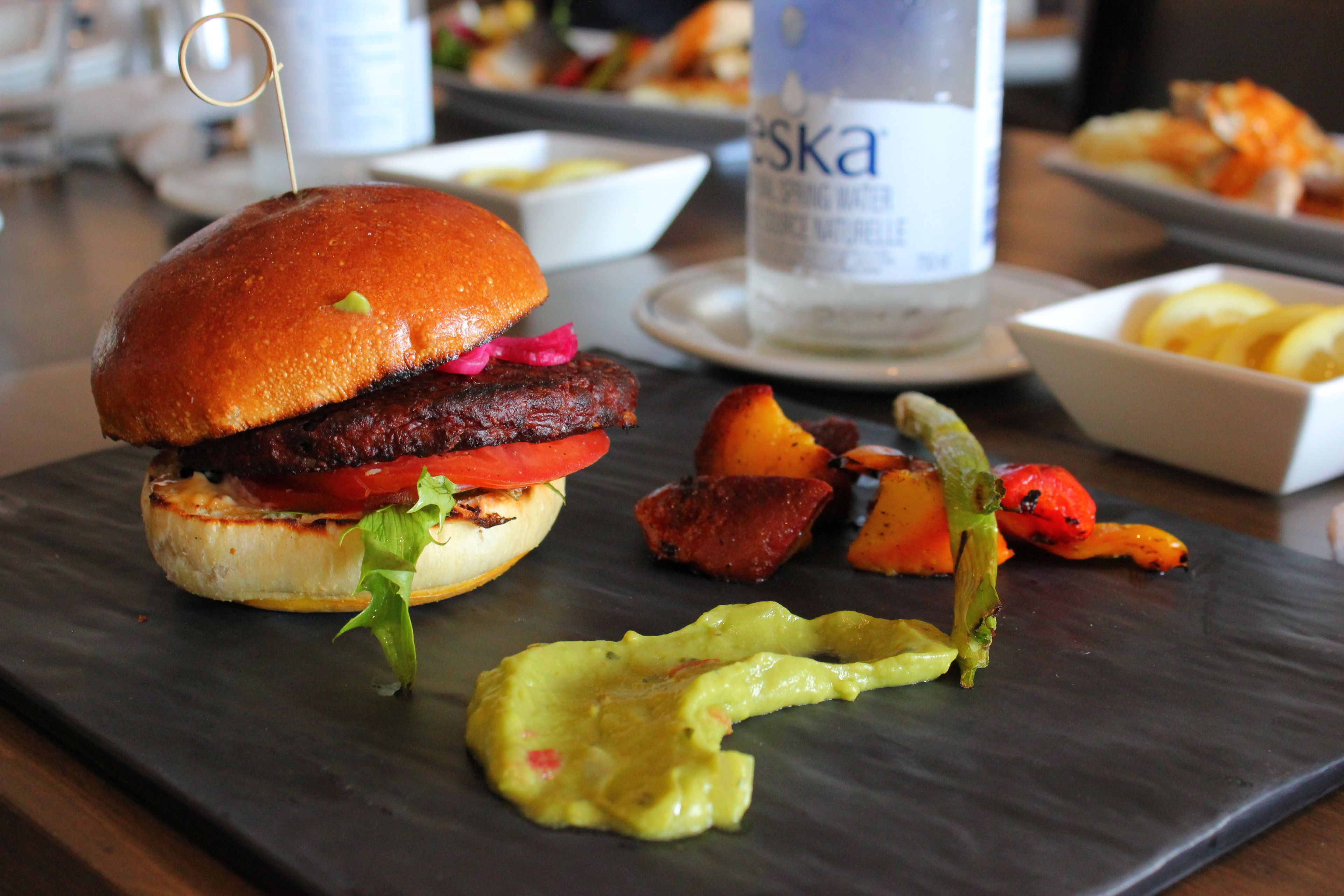 Un repas composé d’un hamburger, de légumes rôtis et d’une portion de sauce, le tout servi sur une assiette en ardoise.