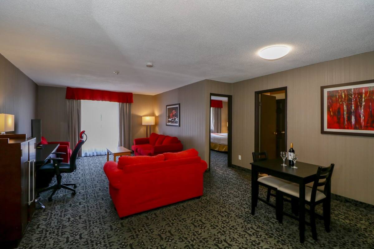 A hotel room with a red sofa, a dining table with chairs, a desk with a chair, a lamp, and a framed picture on the wall.